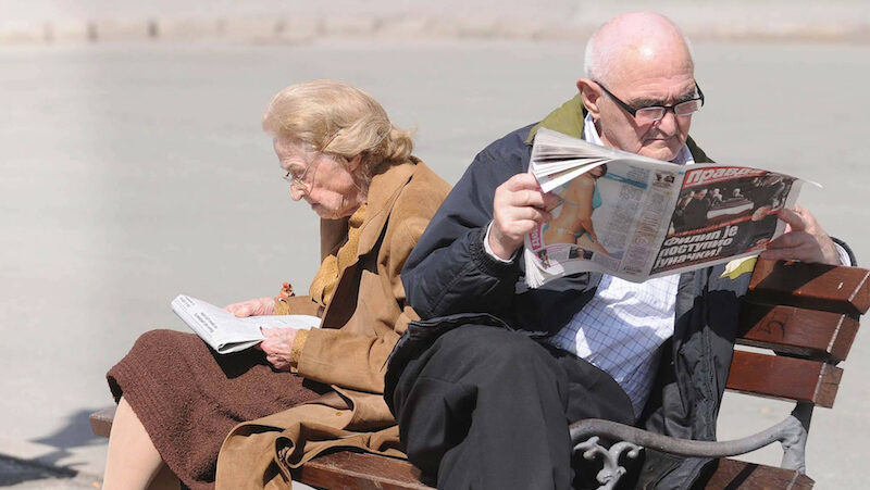 Dva penzionera sede na klupi i citaju novine